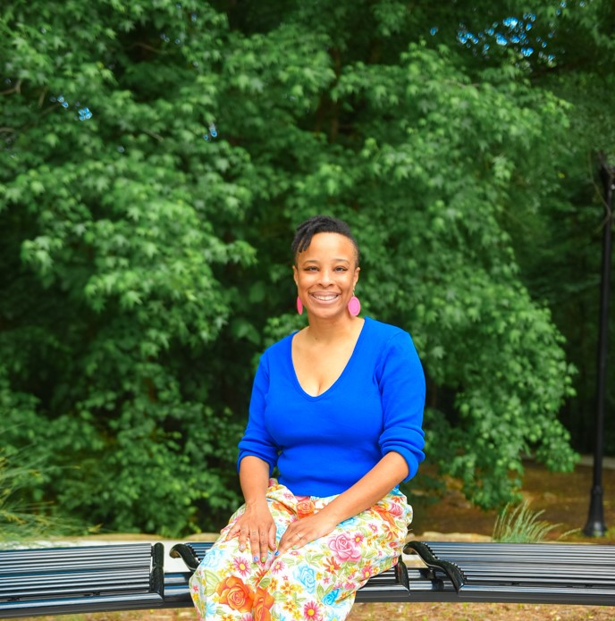 Author Neesha Powell-Ingabire in a blue sweater and pink patterned pants sitting on a park bench.