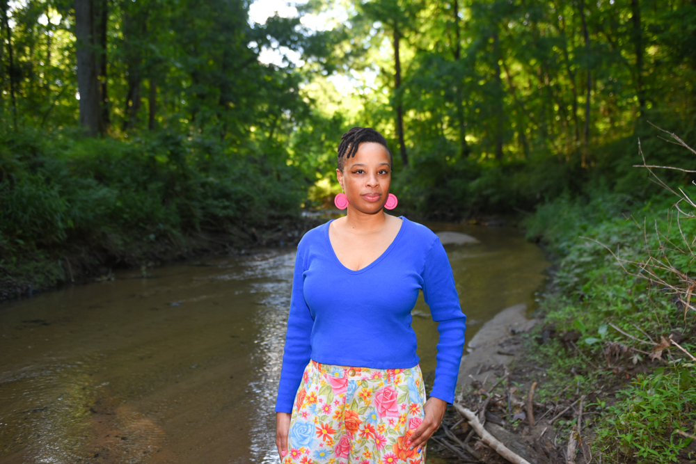 Neesha Powell-Ingabire in a blue shirt with pink-patterned pants and pink, round earrings standing in front of a creek with green grass.