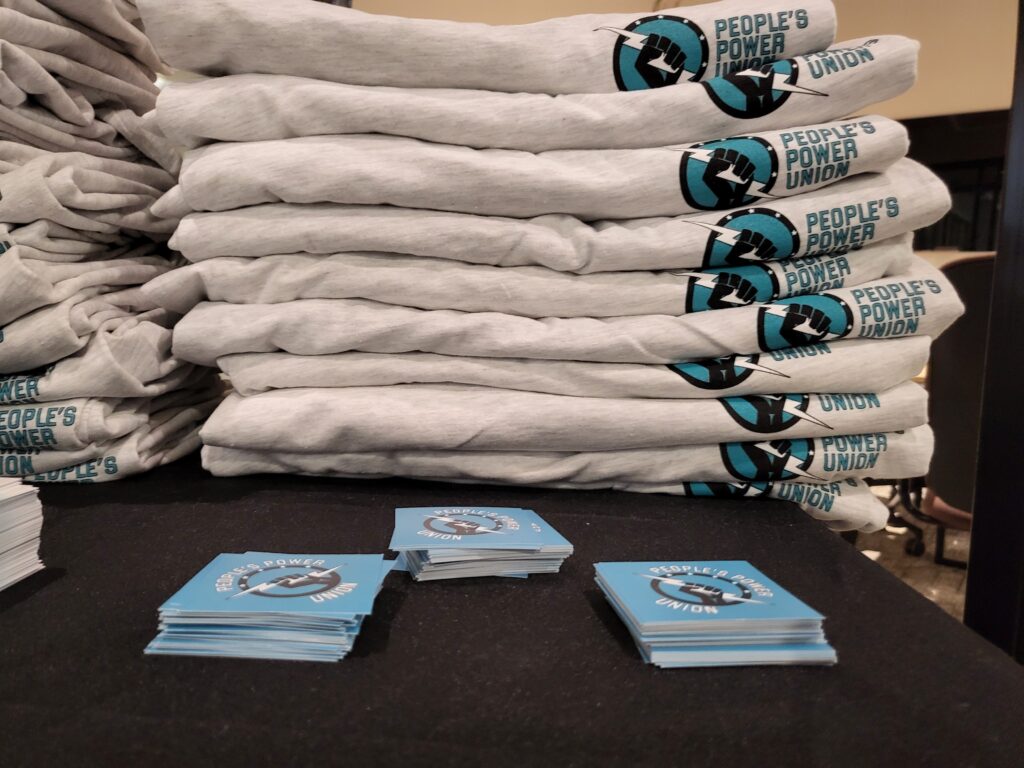 A photo of table with a black tablecloth with folded t-shirts and stickers showing the People's Power Union logo.